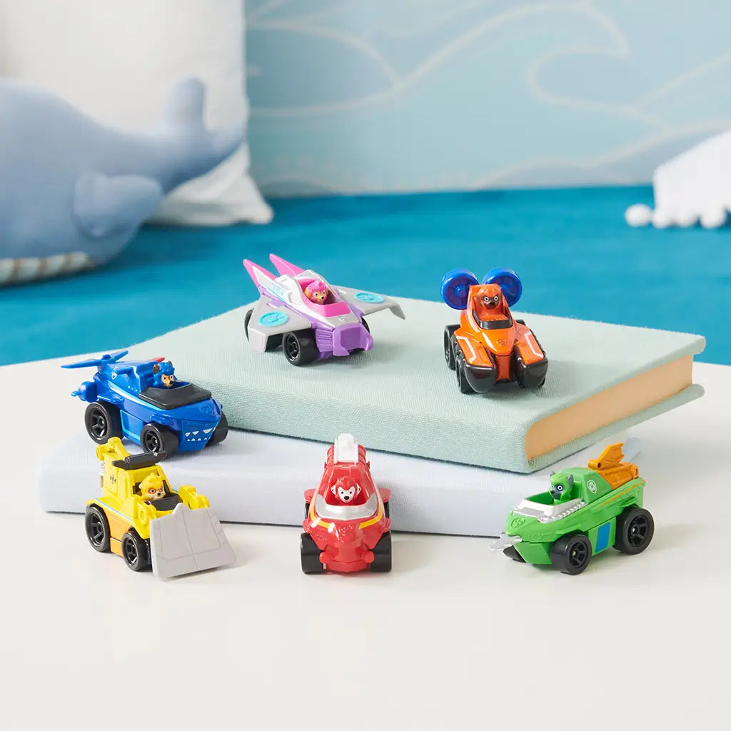 Paw Patrol Aqua Pups toy vehicles in bold plastic colors with smooth glossy texture, small rounded car shapes with character figures, arranged on stacked books in a bright indoor display setting.