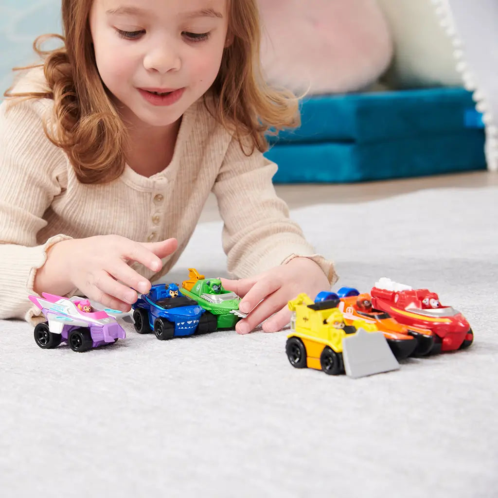PAW Patrol toy cars in vibrant multicolor plastic and metal, small rounded rescue vehicle shapes for preschool play, young child interacting with Zuma and friends on a soft carpet in a bright indoor room