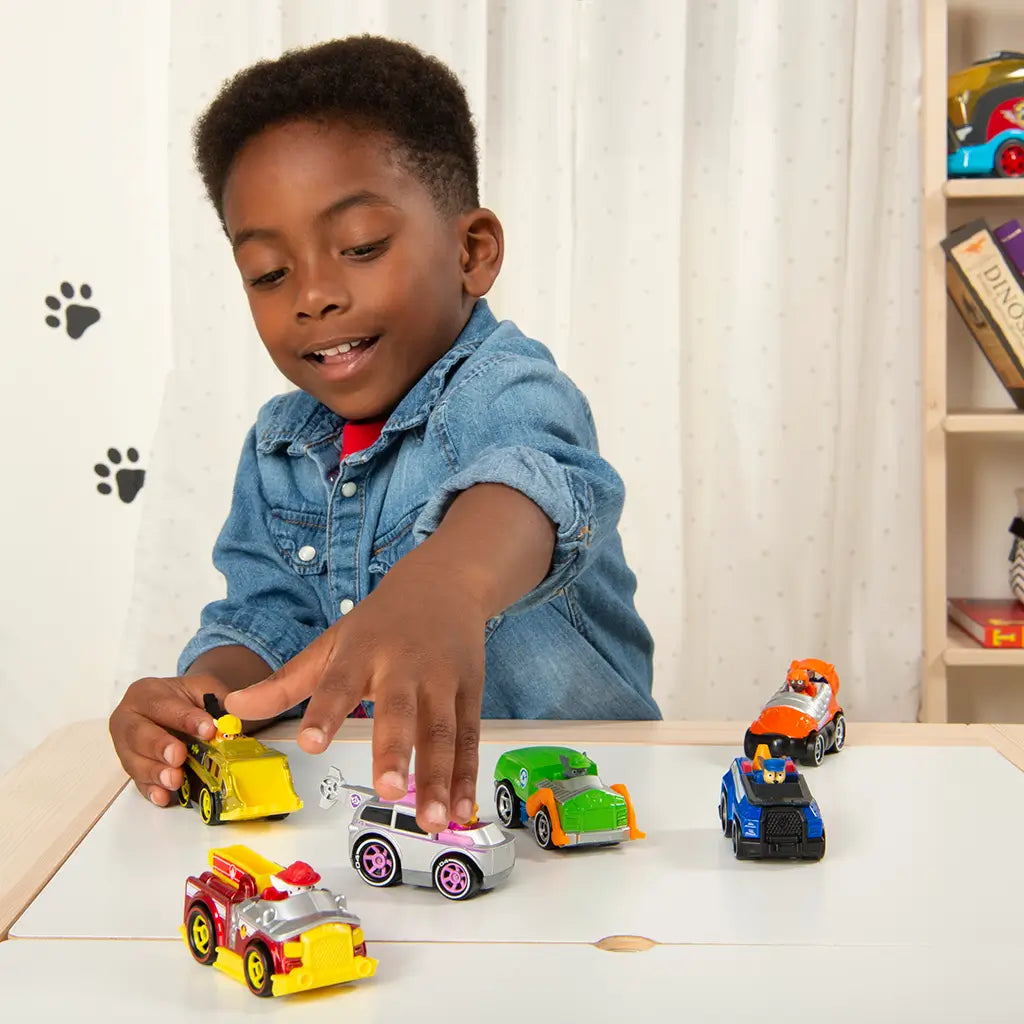 Colorful Paw Patrol toy cars set with red, yellow, green, and blue die-cast vehicles, a young child playing on a tabletop indoors with shelves, books, and toys in the background.