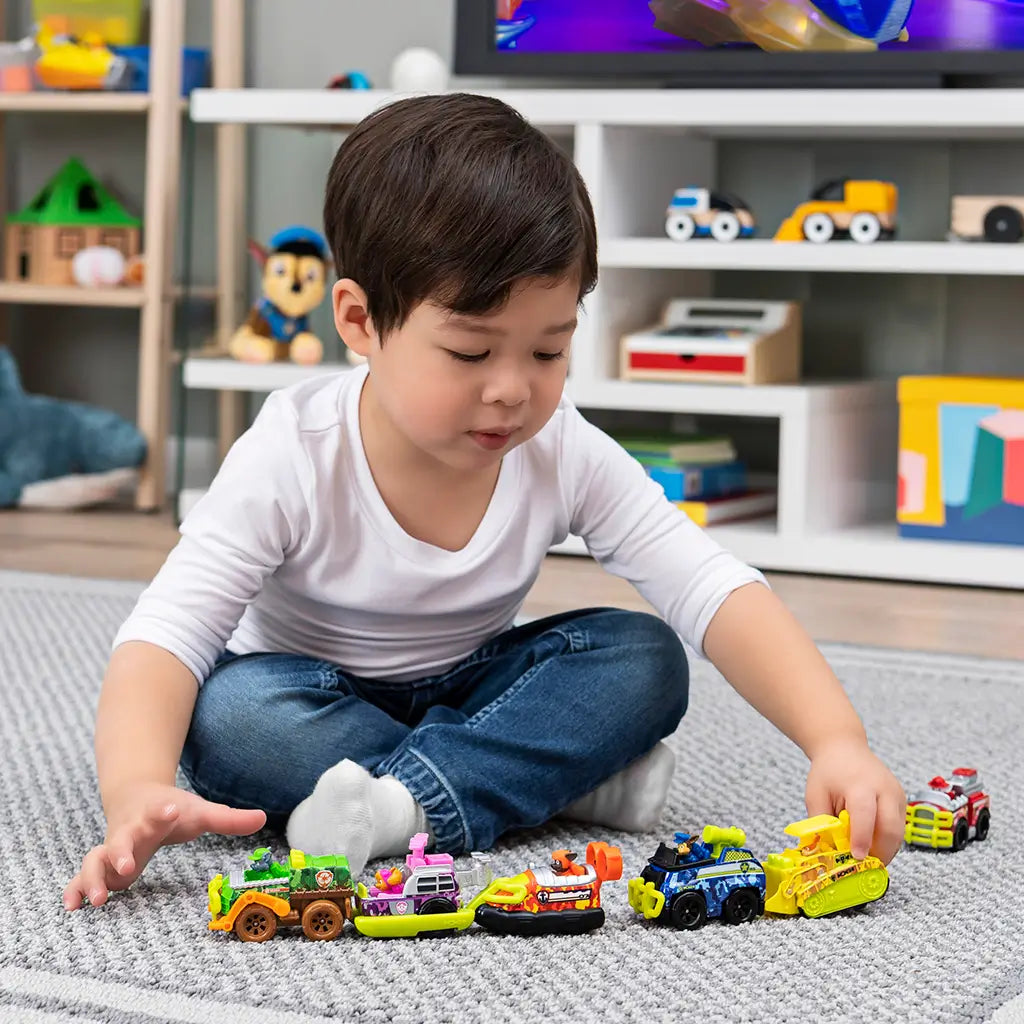 Colorful plastic Paw Patrol toy vehicles arranged in a row as a young child in white shirt and blue jeans sits cross-legged on gray carpet, playing indoors with shelves and toys in background.