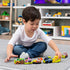 Colorful plastic Paw Patrol toy vehicles arranged in a row as a young child in white shirt and blue jeans sits cross-legged on gray carpet, playing indoors with shelves and toys in background.