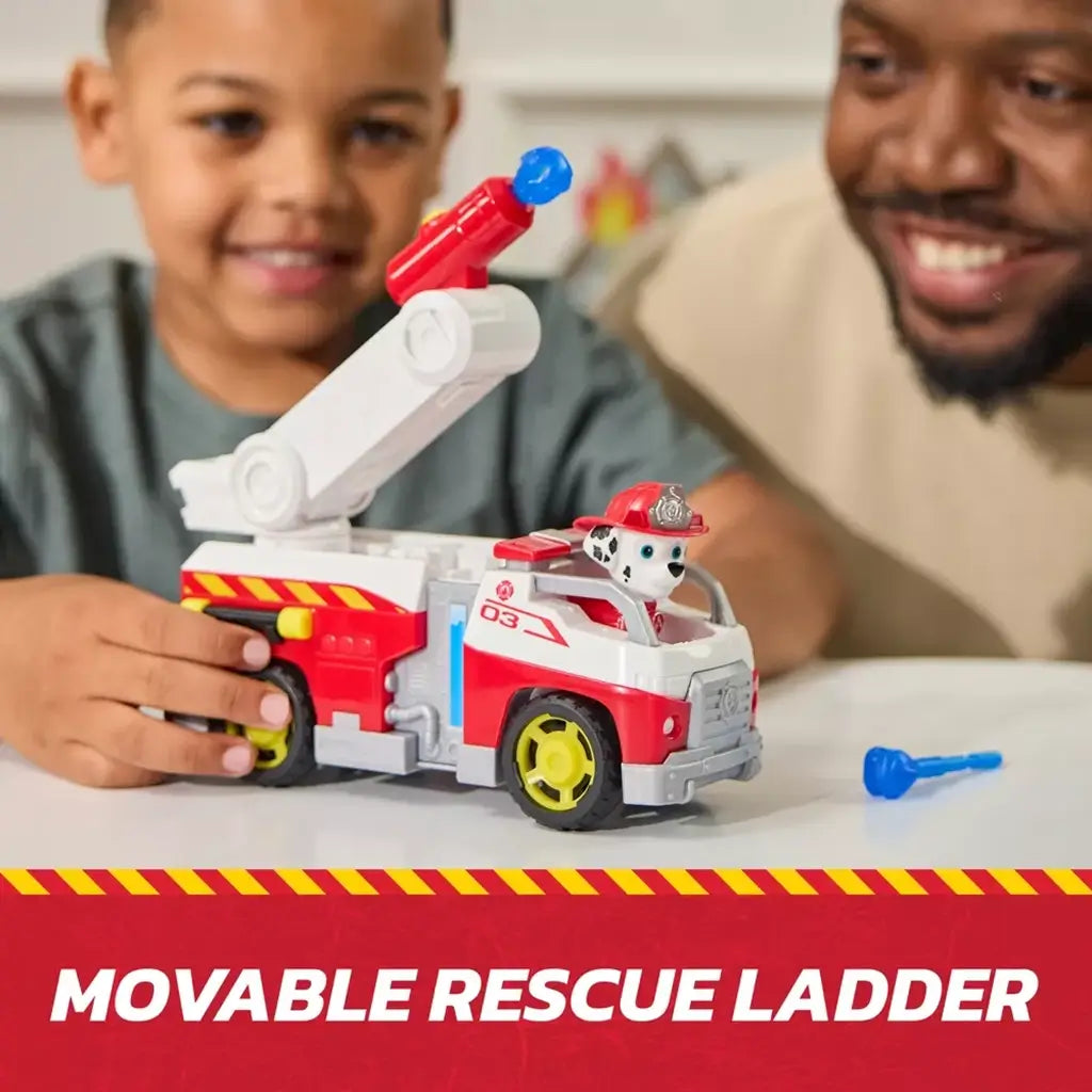 PAW Patrol Marshall fire truck toy in red and white molded plastic with extendable gray ladder and rotating red launcher, shown in a child’s hands on a tabletop during imaginative rescue play.