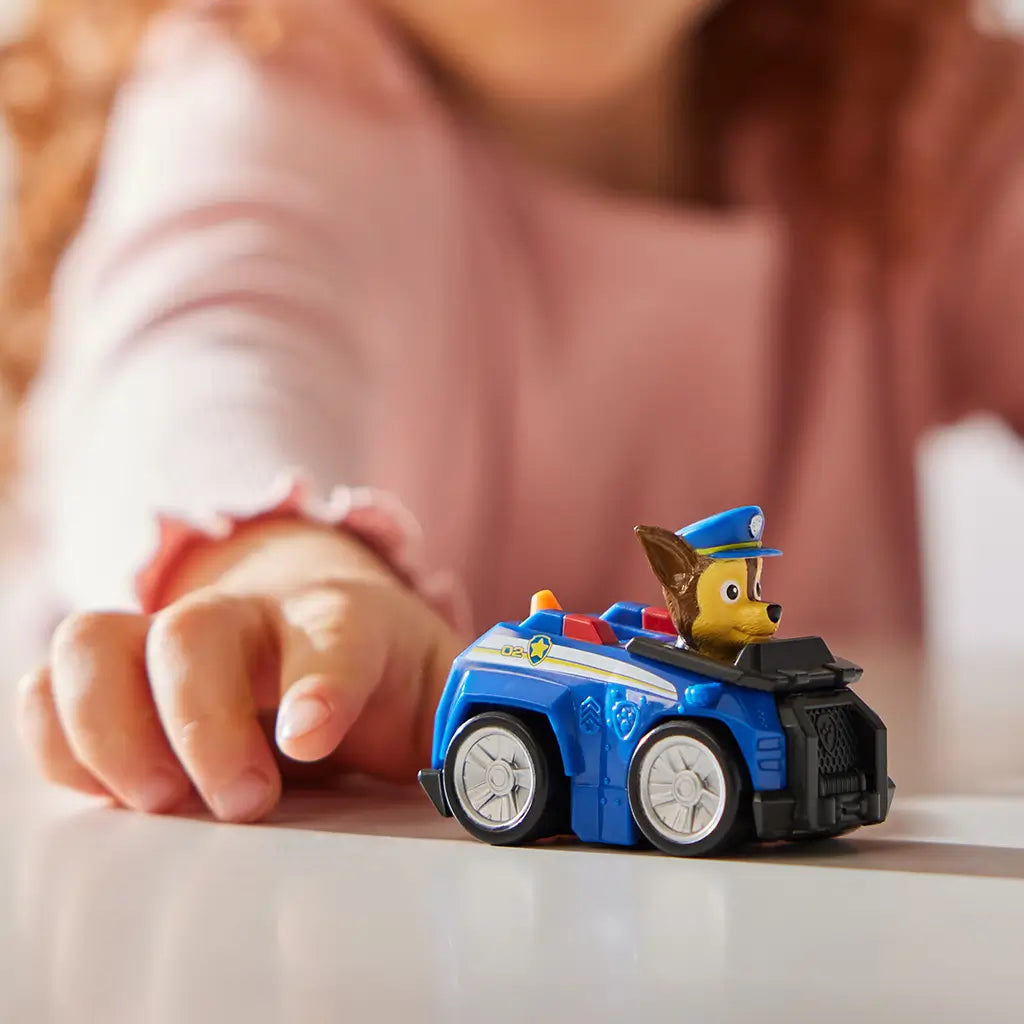 Paw Patrol Chase toy car in blue plastic with black grille, silver wheels, and brown pup figure wearing a blue police hat, compact racer design for kids play, set on a tabletop with a child’s hand in a softly lit indoor background