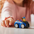 Paw Patrol Chase toy car in blue plastic with black grille, silver wheels, and brown pup figure wearing a blue police hat, compact racer design for kids play, set on a tabletop with a child’s hand in a softly lit indoor background