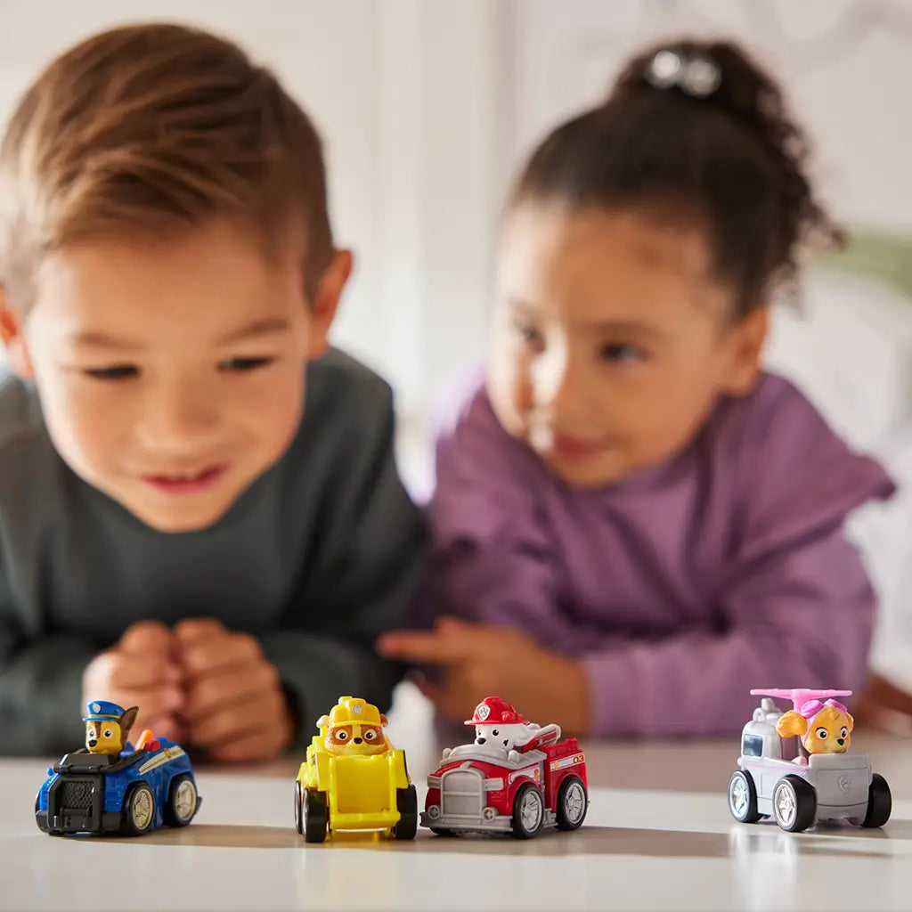 Paw Patrol toy cars in blue, yellow, red, and pink plastic with character figures and smooth rounded racer shapes, designed for kids imaginative play, lined on a tabletop with two children watching in a bright indoor setting
