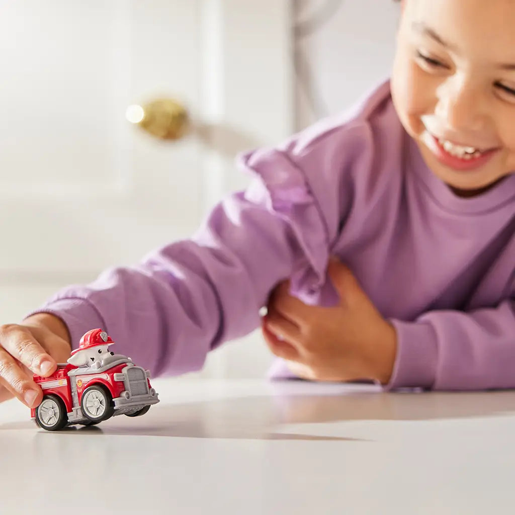 Paw Patrol Marshall toy car in red plastic with silver wheels and white Dalmatian pup wearing firefighter helmet, compact racer design for kids play, shown on tabletop with a smiling child in a softly lit indoor background