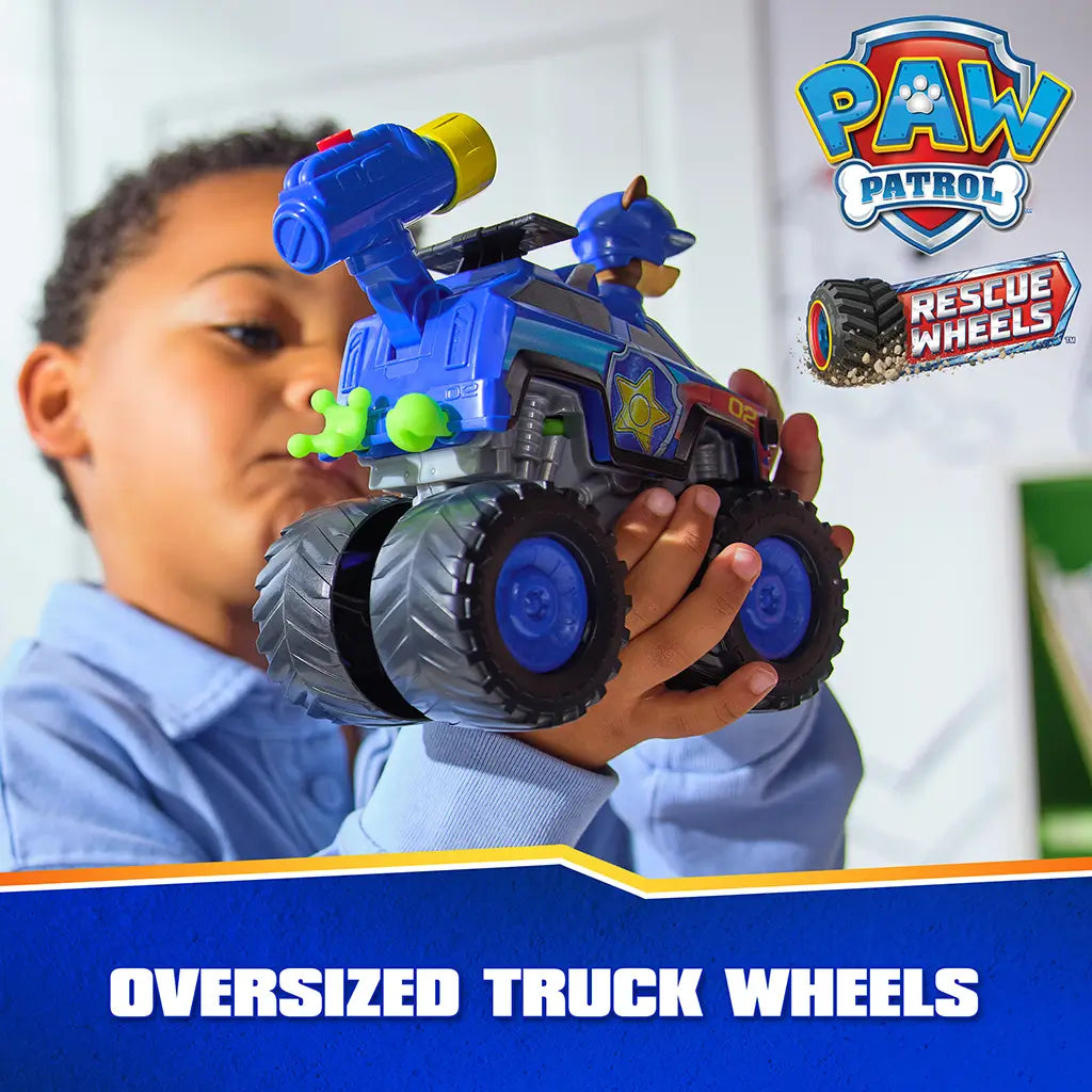 PAW Patrol toy truck blue plastic body with extra large textured black wheels and detailed decals, held by child highlighting oversized tire design in bright indoor playroom environment.