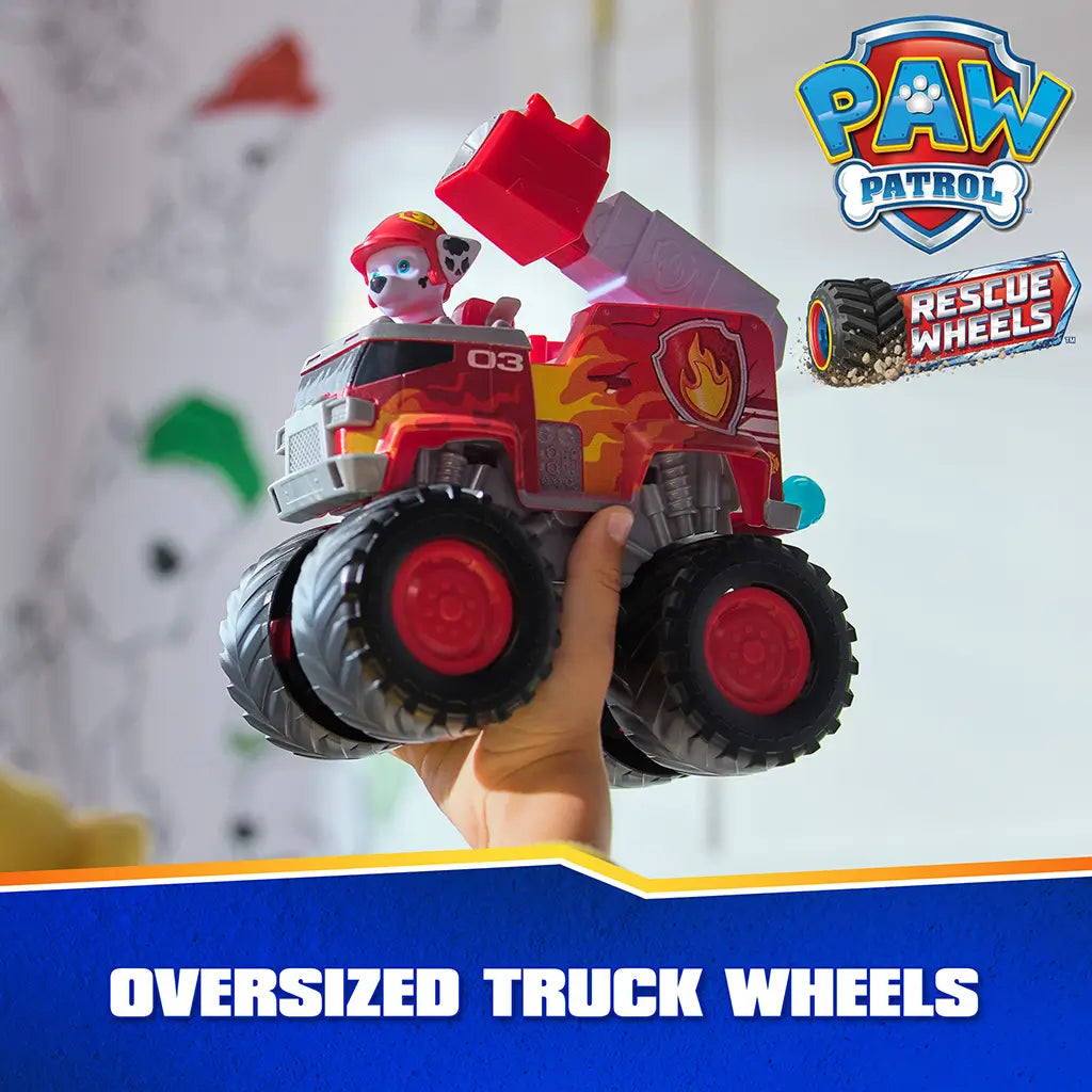 PAW Patrol Marshall fire truck toy displays bold red plastic body with flame decals, large oversized truck wheels, and raised ladder detail, held in hand to highlight durable design in a softly lit indoor background.