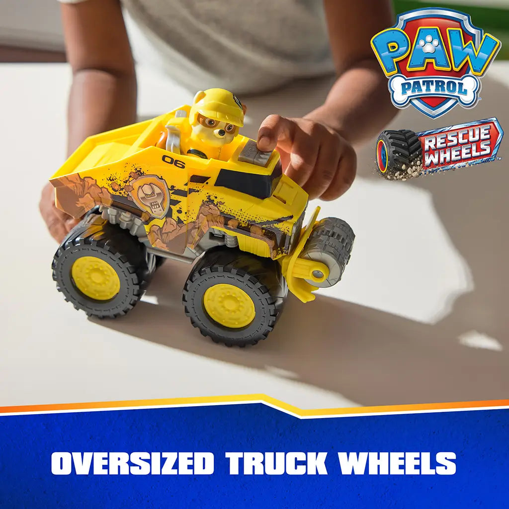 PAW Patrol Rubble truck toy shows bright yellow plastic body with textured mud graphics, large rubber-like wheels, and front roller attachment, demonstrated in a child’s hands on a tabletop play setting.