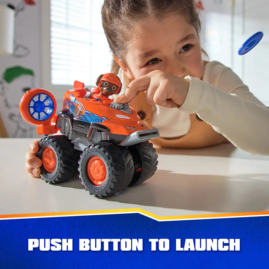 PAW Patrol Zuma hovercraft toy features bright orange plastic body with glossy and textured surfaces, large rugged wheels, and push-button launcher, demonstrated by a child during interactive tabletop play.