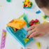 Play-Doh blue plastic extruder with orange lever and molded shape cavities surrounded by bright dough pieces as child’s hand presses tool on white table during creative play activity.