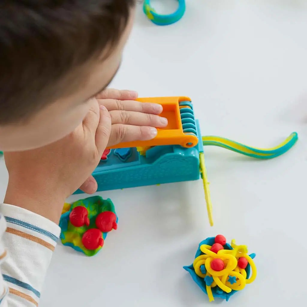 Play-Doh blue and orange extruder producing long yellow and green dough strands as child presses lever on white tabletop with colorful molded pieces scattered around the scene.