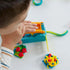 Play-Doh blue and orange extruder producing long yellow and green dough strands as child presses lever on white tabletop with colorful molded pieces scattered around the scene.