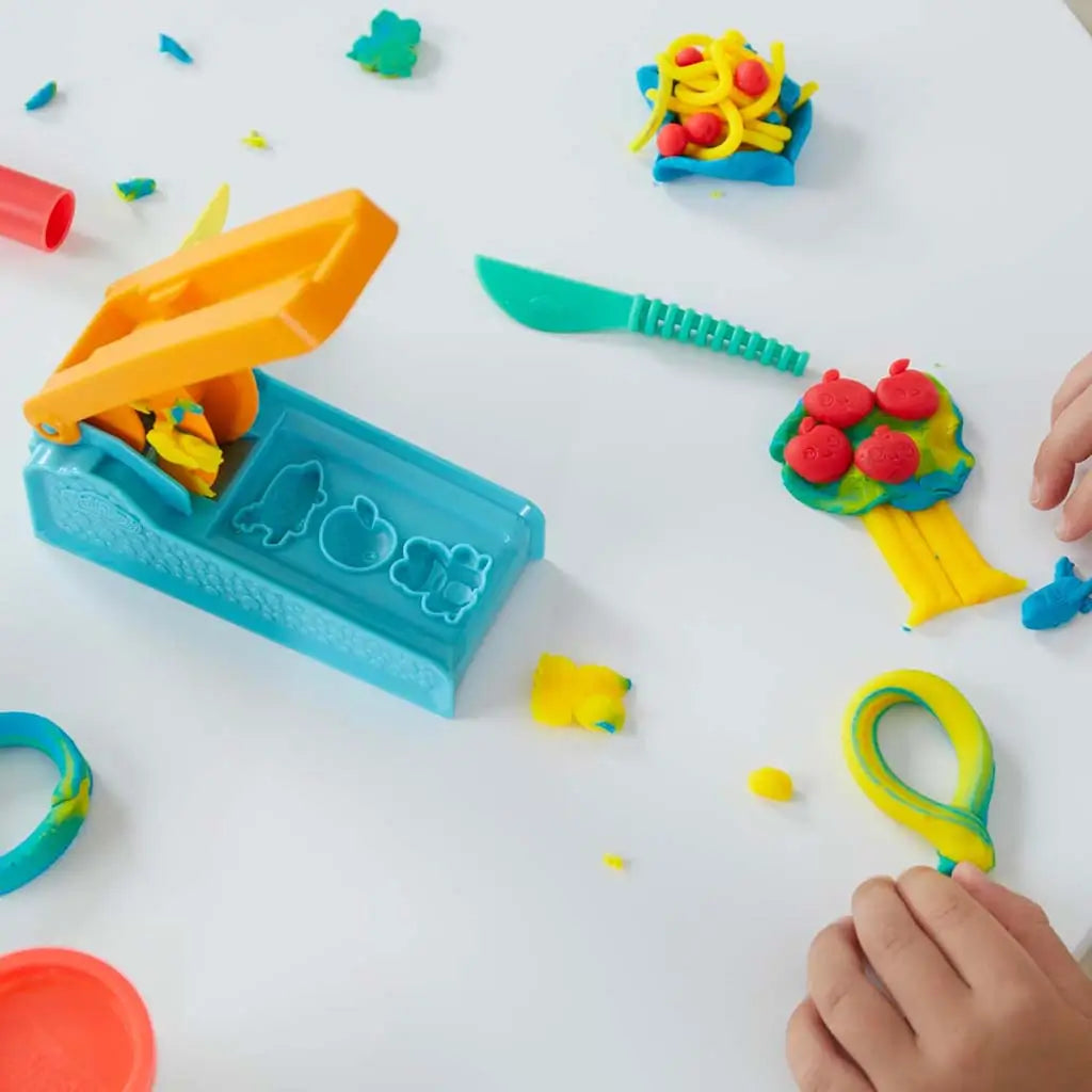 Play-Doh blue extruder with orange handle displayed on white table among yellow, red and blue dough shapes including tree and noodles near plastic knife during hands-on creative activity.