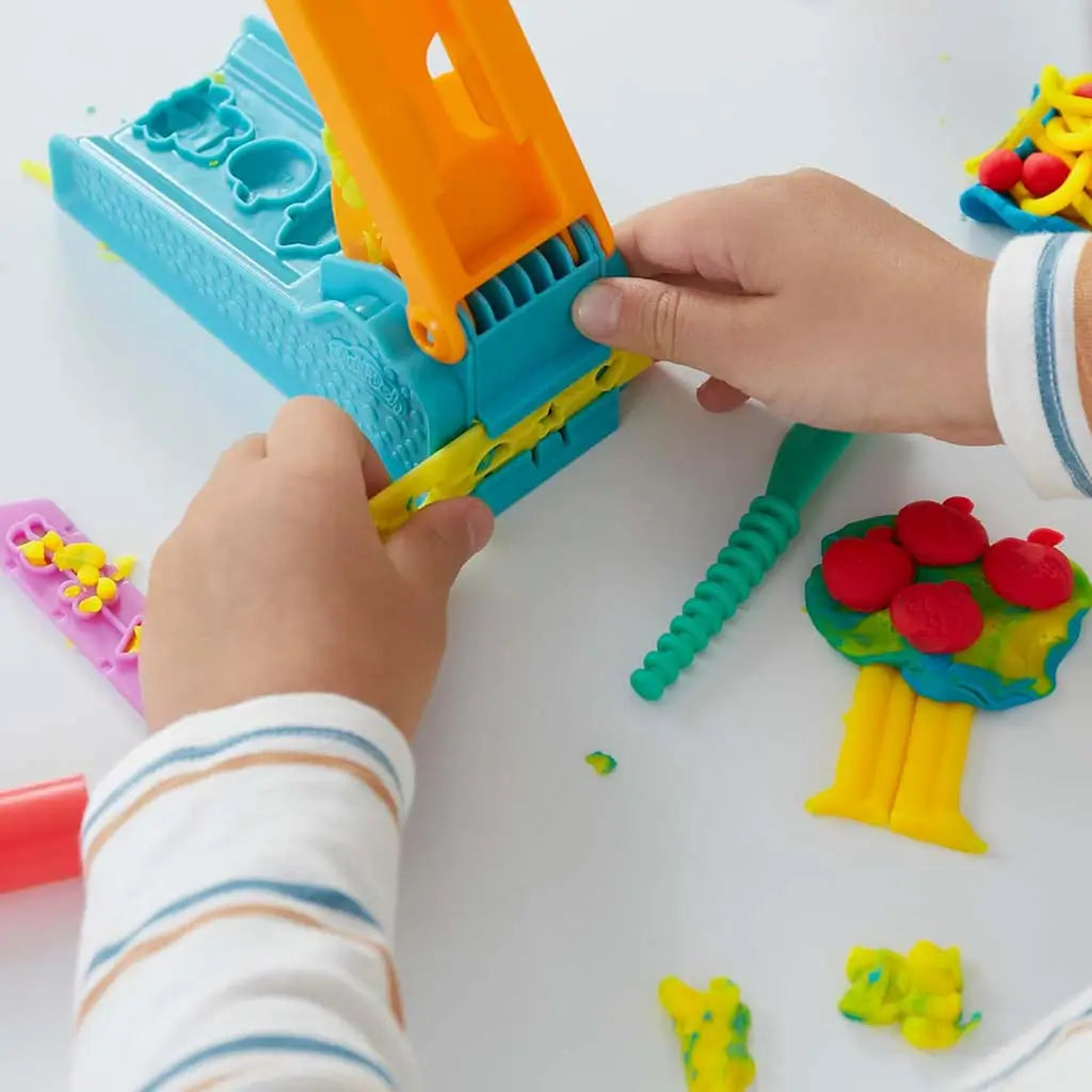 Play-Doh blue extruder with orange compression bar forming star shapes from yellow dough as child’s hands guide strip on white table surrounded by plastic knife and bright molded pieces.