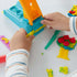 Play-Doh blue extruder with orange compression bar forming star shapes from yellow dough as child’s hands guide strip on white table surrounded by plastic knife and bright molded pieces.