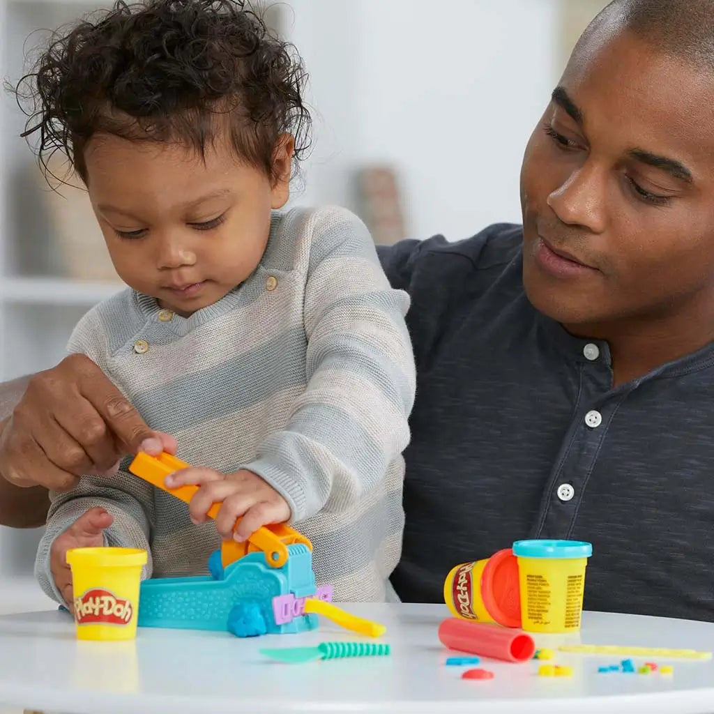 Play-Doh blue extruder with orange lever used by toddler guided by adult at small white table with open yellow dough tub nearby during shared creative play session.