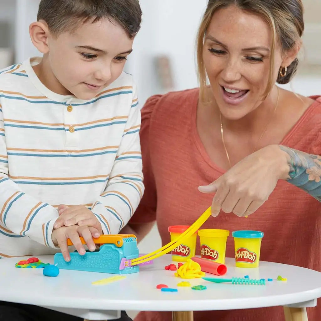 Play-Doh blue extruder producing long yellow dough ribbon as child presses lever while adult points beside three bright tubs on small white table during interactive crafting moment.