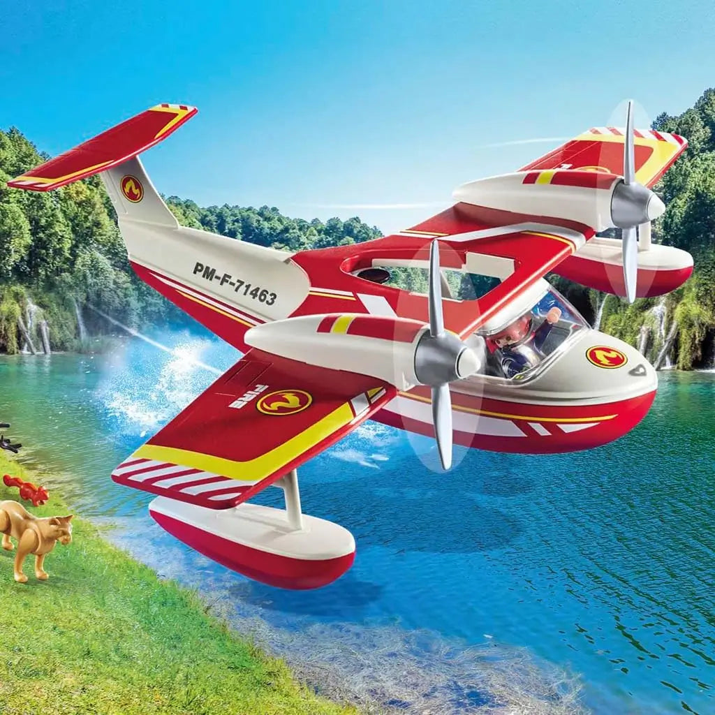 Playmobil seaplane banking low over turquoise water near waterfalls, with animal figure on grassy shore and bright flame pieces around a scorched tree illustrating rescue action.