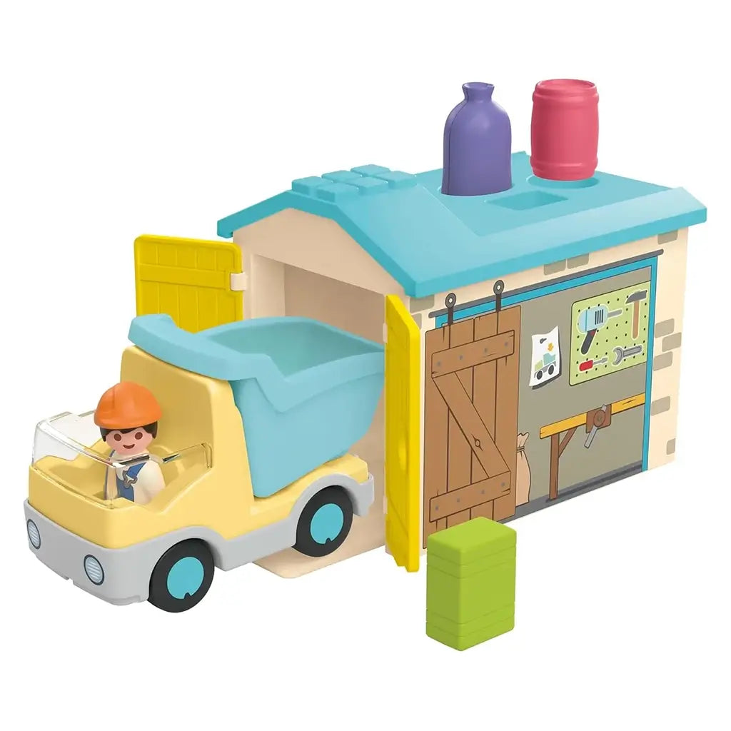 Playmobil Junior garage view shows blue roof with shape-sorting holes, sliding barn-style door and pastel dump truck exiting while colorful pieces sit ready for early construction learning.