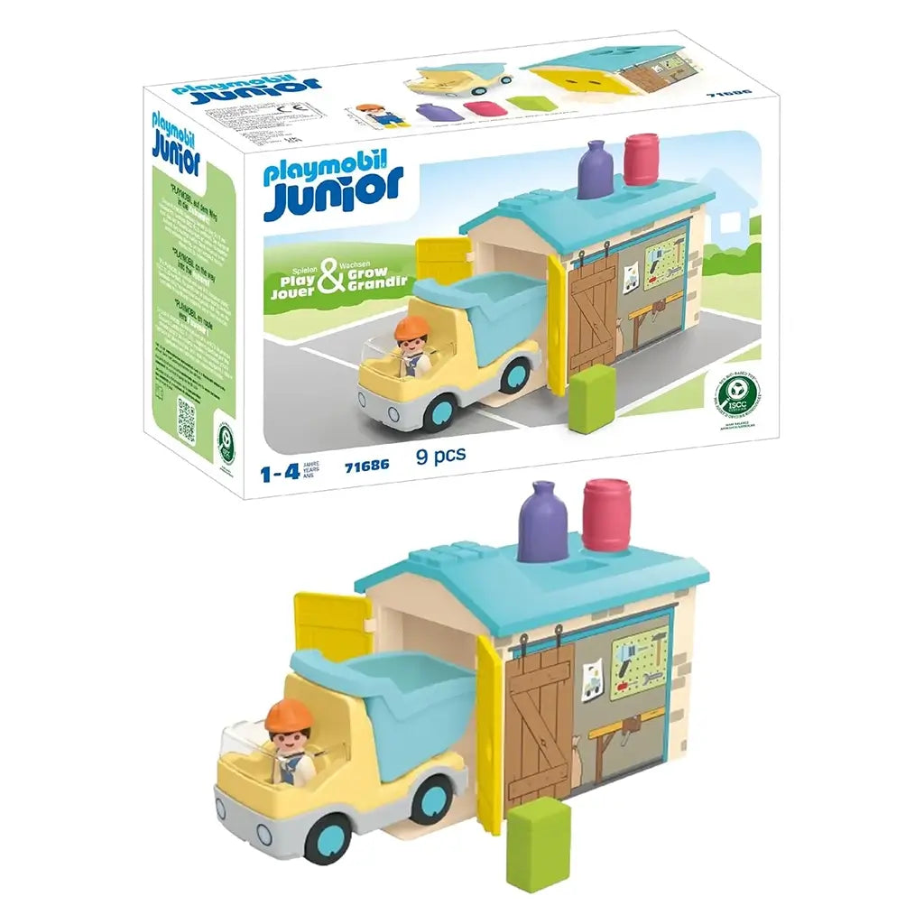 Playmobil Junior construction toy shows bright plastic truck with blue dump bed beside a small garage with sliding doors, tool graphics and shape-sorting roof in a simple outdoor scene.