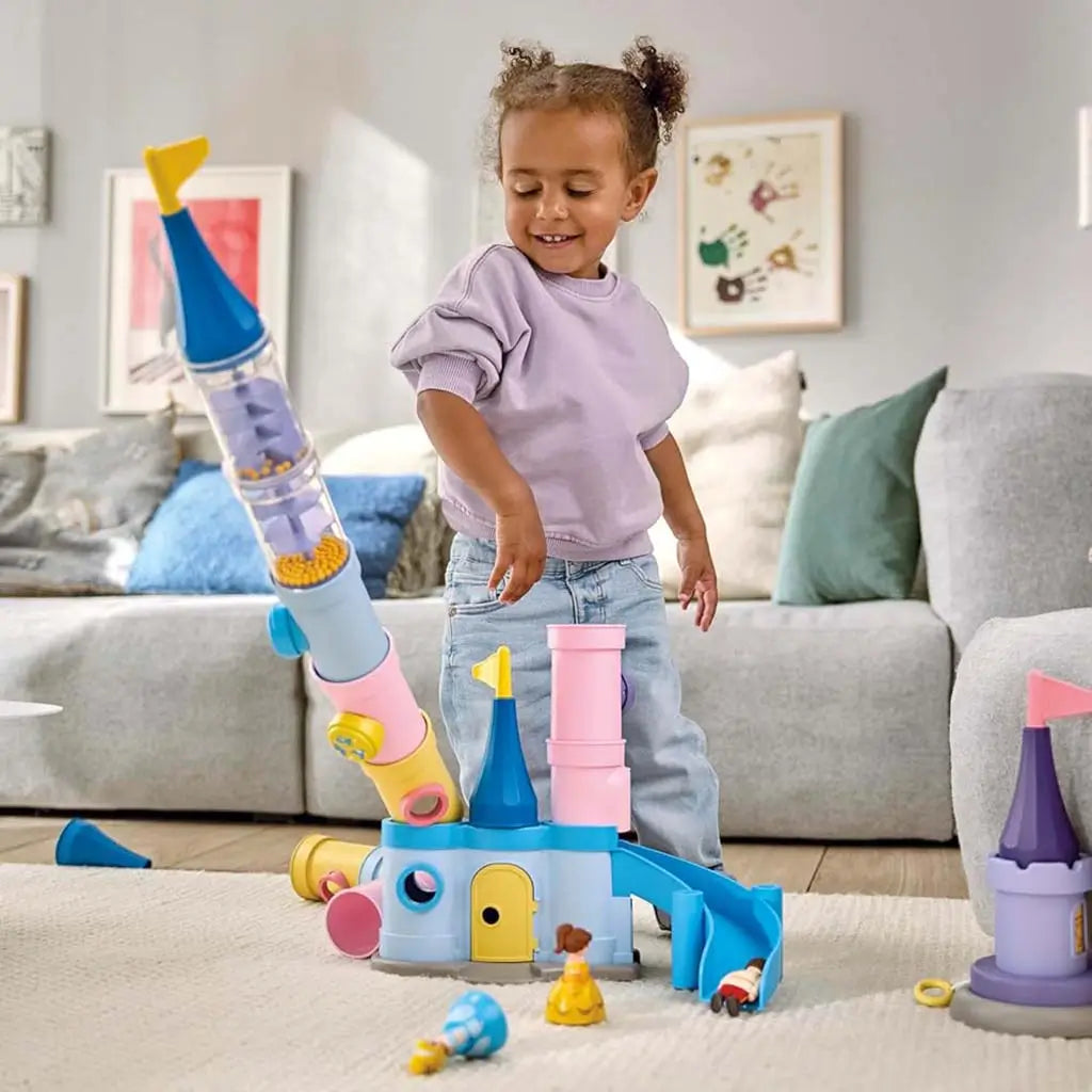 A young child stands beside a colorful Playmobil Disney castle playset built from pastel plastic tubes with spiral-ramp pieces and yellow beads, showing imaginative stacking play in a bright living room.