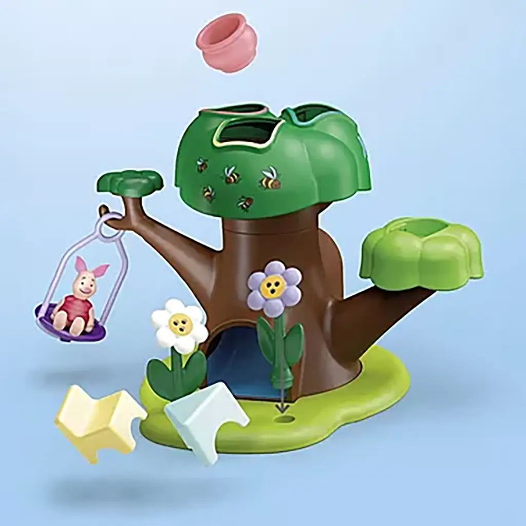 Disney Playmobil Junior Winnie the Pooh tree house toy with green canopy, brown trunk, bright flower pieces, swing holding Piglet, and colorful shape blocks shown in motion against a light blue backdrop.