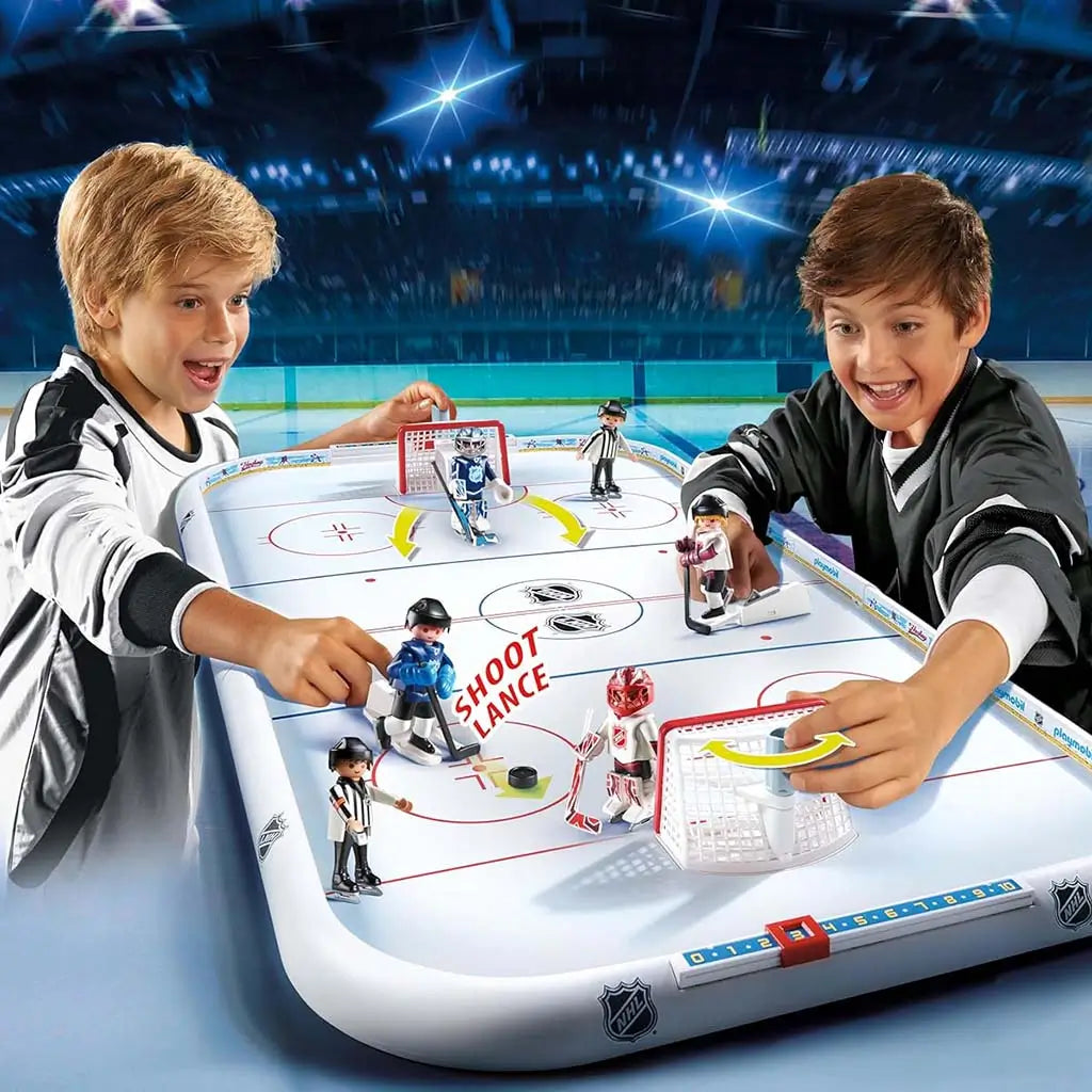 Playmobil NHL tabletop hockey game with glossy white rink and movable plastic players being controlled by two excited children seated at the game table in a stadium‑style background.