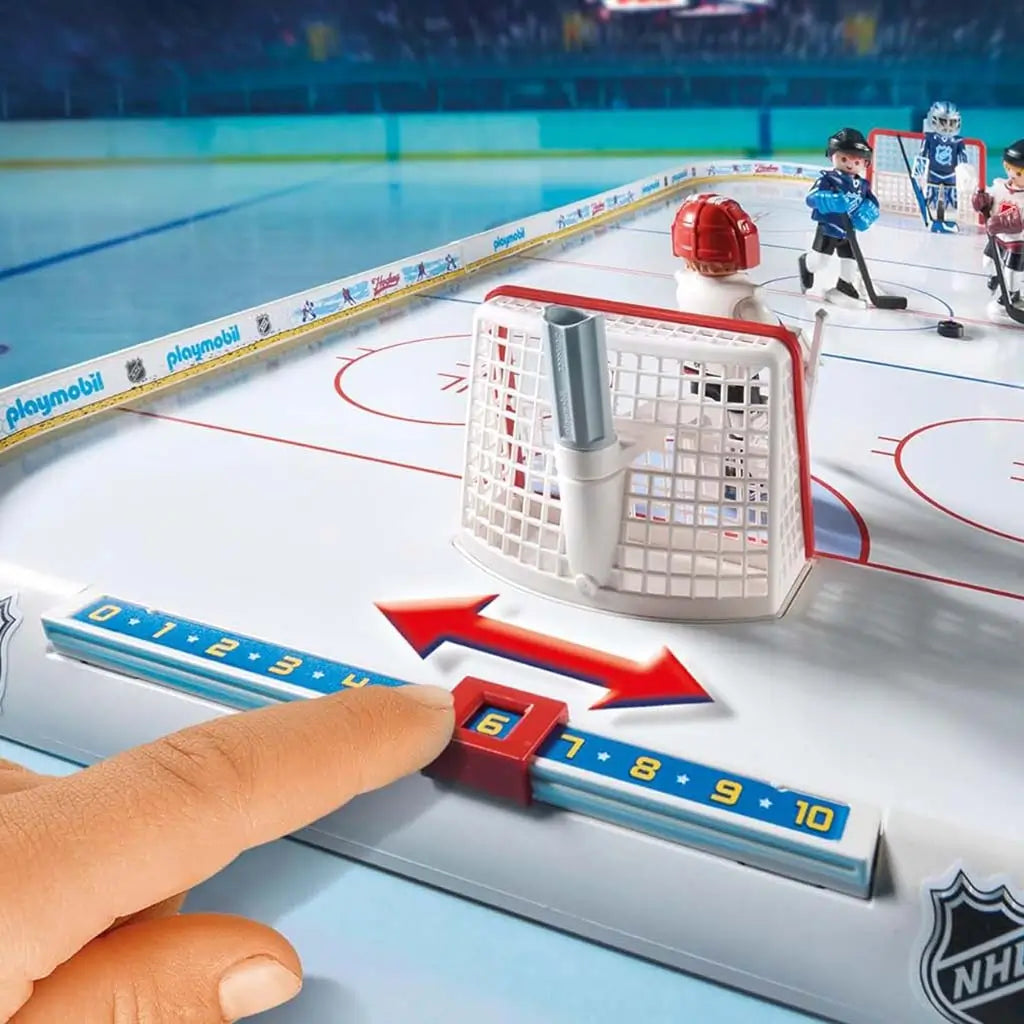 Playmobil NHL tabletop rink close‑up shows white goal net, sliding blue score tracker, smooth ice surface markings, and player figures positioned in a brightly lit stadium-style environment.