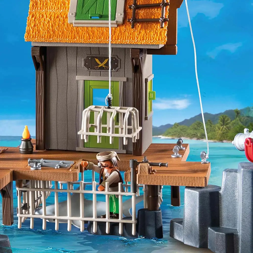 Close-up of the Playmobil pirate shack features a working drop cage, wooden deck, and a pirate figure near turquoise water, emphasizing interactive capture play on the tropical pier.