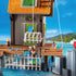 Close-up of the Playmobil pirate shack features a working drop cage, wooden deck, and a pirate figure near turquoise water, emphasizing interactive capture play on the tropical pier.