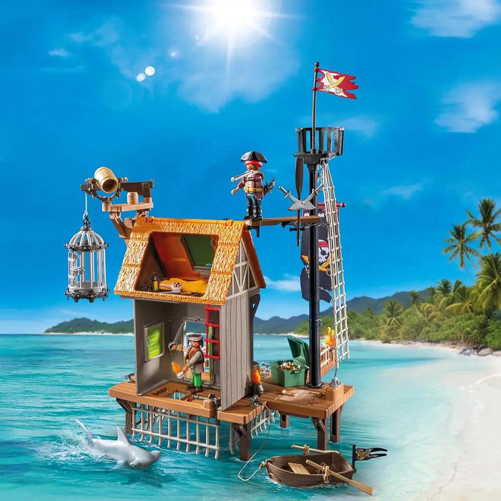 Wide Playmobil Pirates harbor scene with wooden hut, docks, shark, rowboat, flags, and lanterns displayed against a bright tropical beach backdrop, showcasing the full island hideout build.