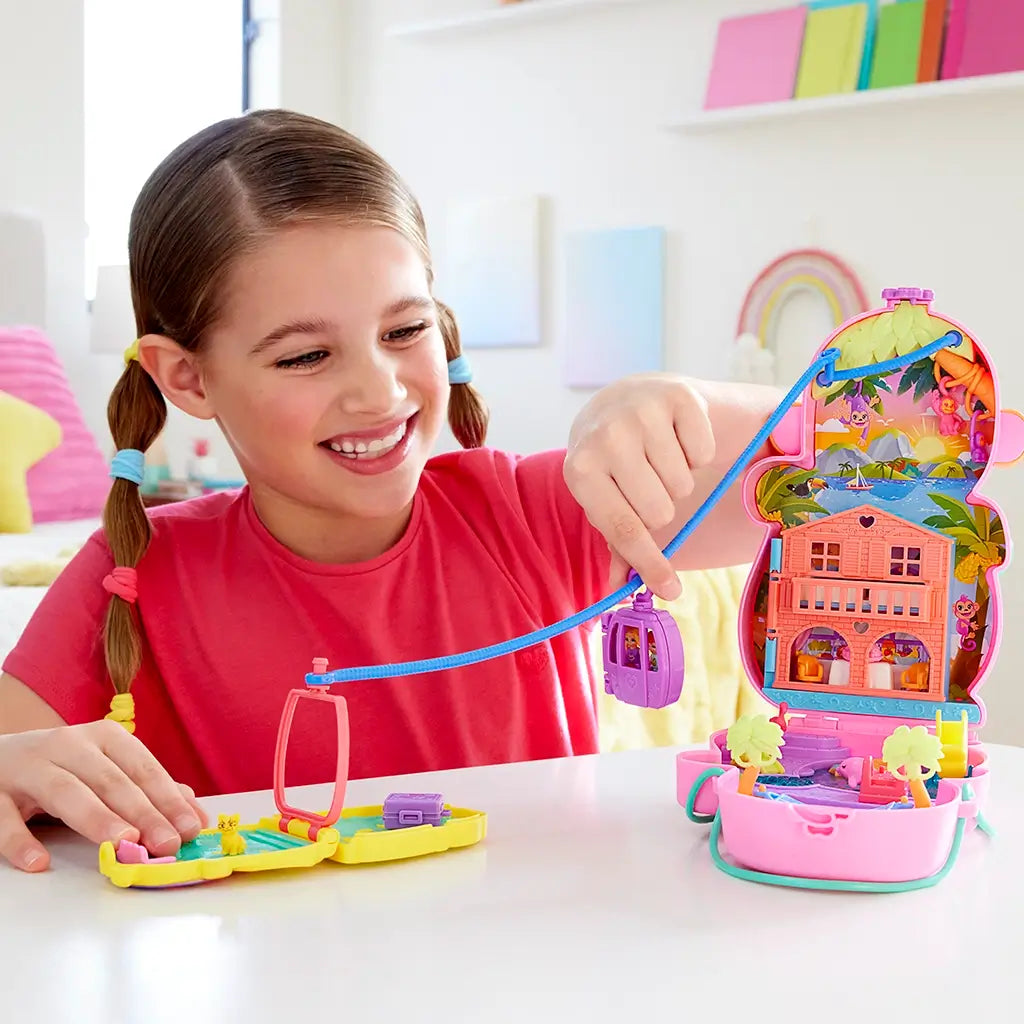 Smiling young girl in pink shirt plays with Polly Pocket Momma Monkey playset, using the zipline gondola feature to connect two colorful compacts on a white table.