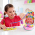 Smiling young girl in pink shirt plays with Polly Pocket Momma Monkey playset, using the zipline gondola feature to connect two colorful compacts on a white table.
