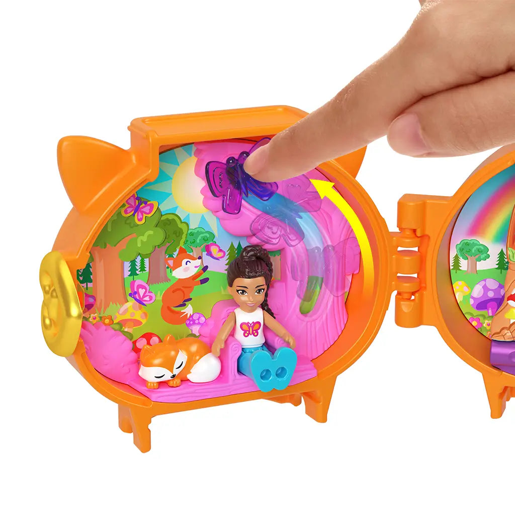 Child’s hand interacting with Polly Pocket fox locket playset, pressing butterfly feature as doll and fox figure rest inside detailed woodland scene with trees, flowers, and colorful designs.