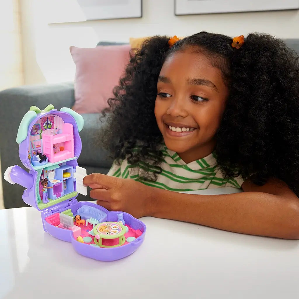 Smiling child plays with Polly Pocket Sleepover Puppy compact, opening dollhouse rooms with tiny dolls, dogs, furniture, and accessories on a table.