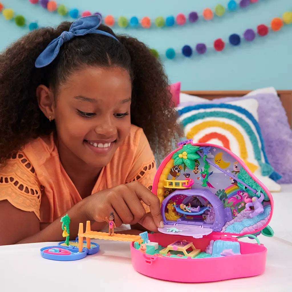 Smiling child in orange shirt plays with Polly Pocket Sloth Family 2-in-1 compact playset, showcasing colorful miniature jungle scene with dolls, bridge, and animal accessories.