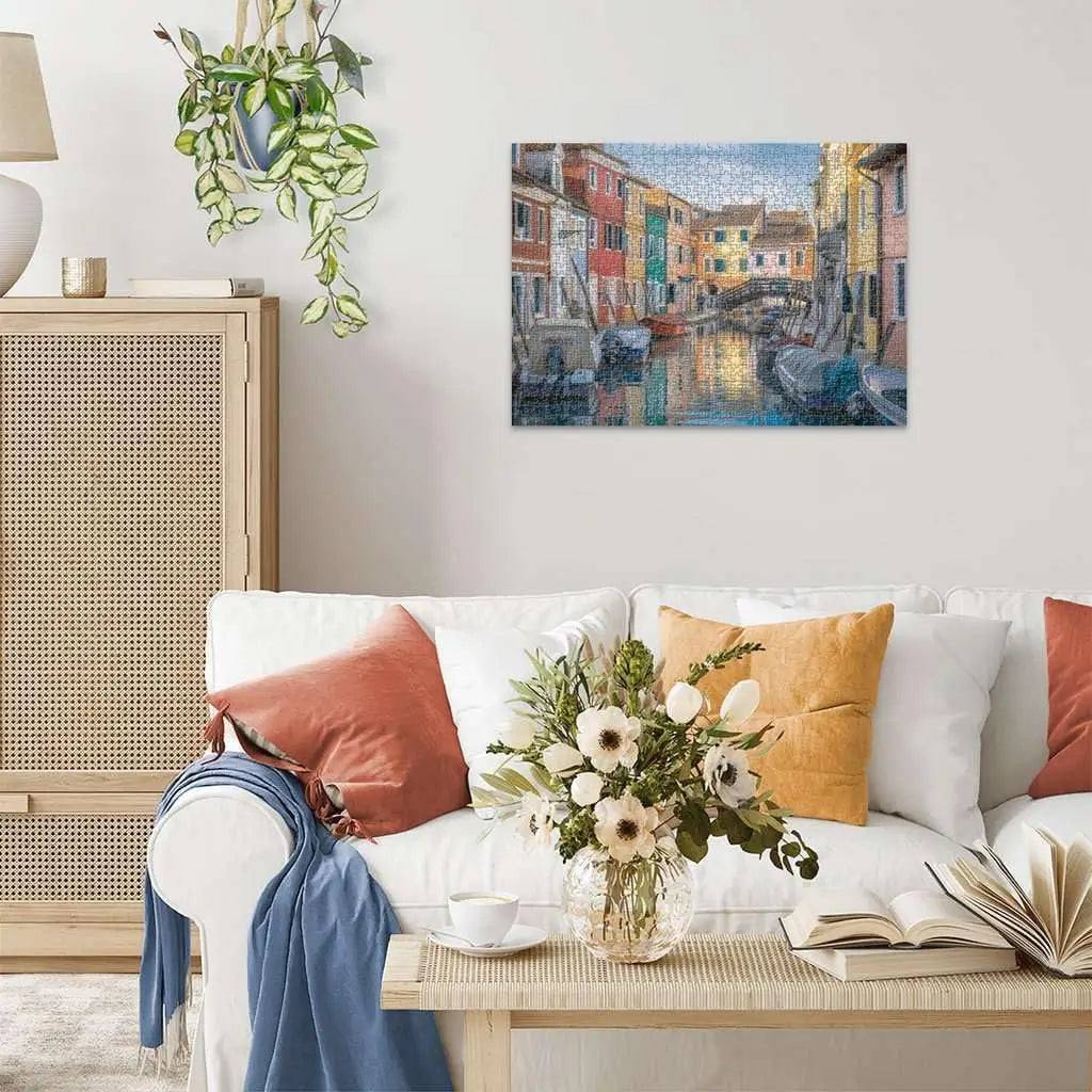 Completed Ravensburger Burano Italy 1000 piece puzzle displayed on wall above white sofa with colorful pillows and flowers, showing vibrant canal cityscape.