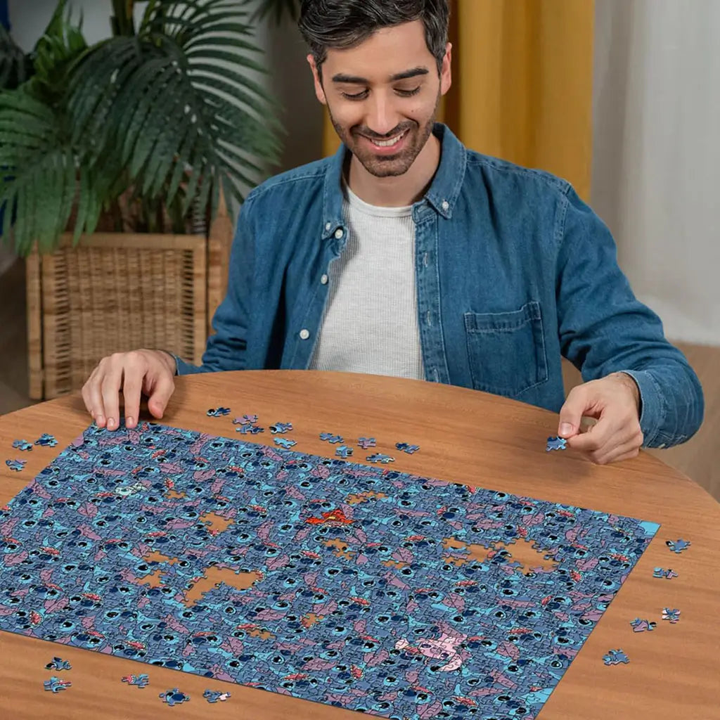 A man in a denim shirt smiles while assembling the Ravensburger Disney Stitch Challenge puzzle on a wooden table, filled with hundreds of expressive Stitch character faces.