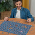 A man in a denim shirt smiles while assembling the Ravensburger Disney Stitch Challenge puzzle on a wooden table, filled with hundreds of expressive Stitch character faces.