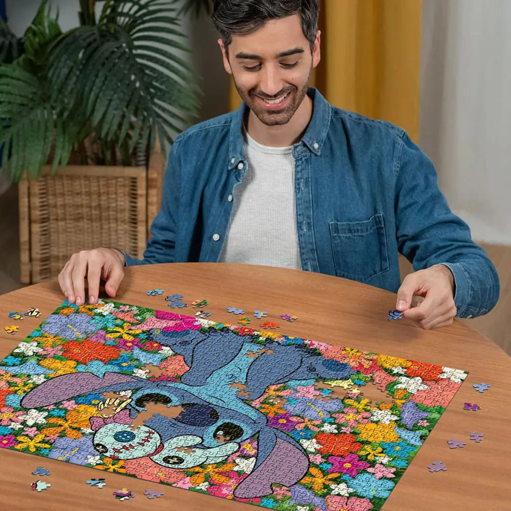 A smiling man in a denim shirt assembles a colorful Ravensburger Disney Stitch 1000-piece puzzle featuring Stitch surrounded by vibrant tropical flowers on a wooden table.