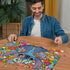 A smiling man in a denim shirt assembles a colorful Ravensburger Disney Stitch 1000-piece puzzle featuring Stitch surrounded by vibrant tropical flowers on a wooden table.