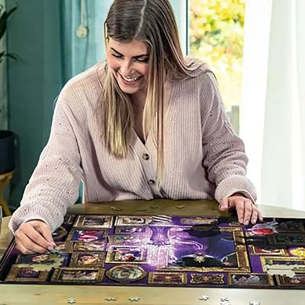 Smiling woman assembling Ravensburger Disney Villainous Evil Queen puzzle on a wooden table, working on detailed purple and gold artwork filled with framed Snow White characters.