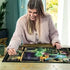 Smiling woman in a cozy sweater assembling Ravensburger Disney Villainous Maleficent puzzle on wooden table, featuring dark green artwork with magical frames and Maleficent’s figure.