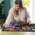 Smiling woman assembles Ravensburger Disney Villainous Ursula puzzle at a wooden table, surrounded by scattered pieces, highlighting vibrant purple artwork and golden frames.