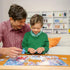 Smiling father and young son sit at a wooden table working on a Ravensburger Encanto puzzle, bonding while assembling colorful pieces in a cozy living room.