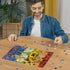 A smiling man in a denim shirt sits at a wooden table assembling the Ravensburger Floral Reflections puzzle, showing colorful flower patterns with scattered pieces.