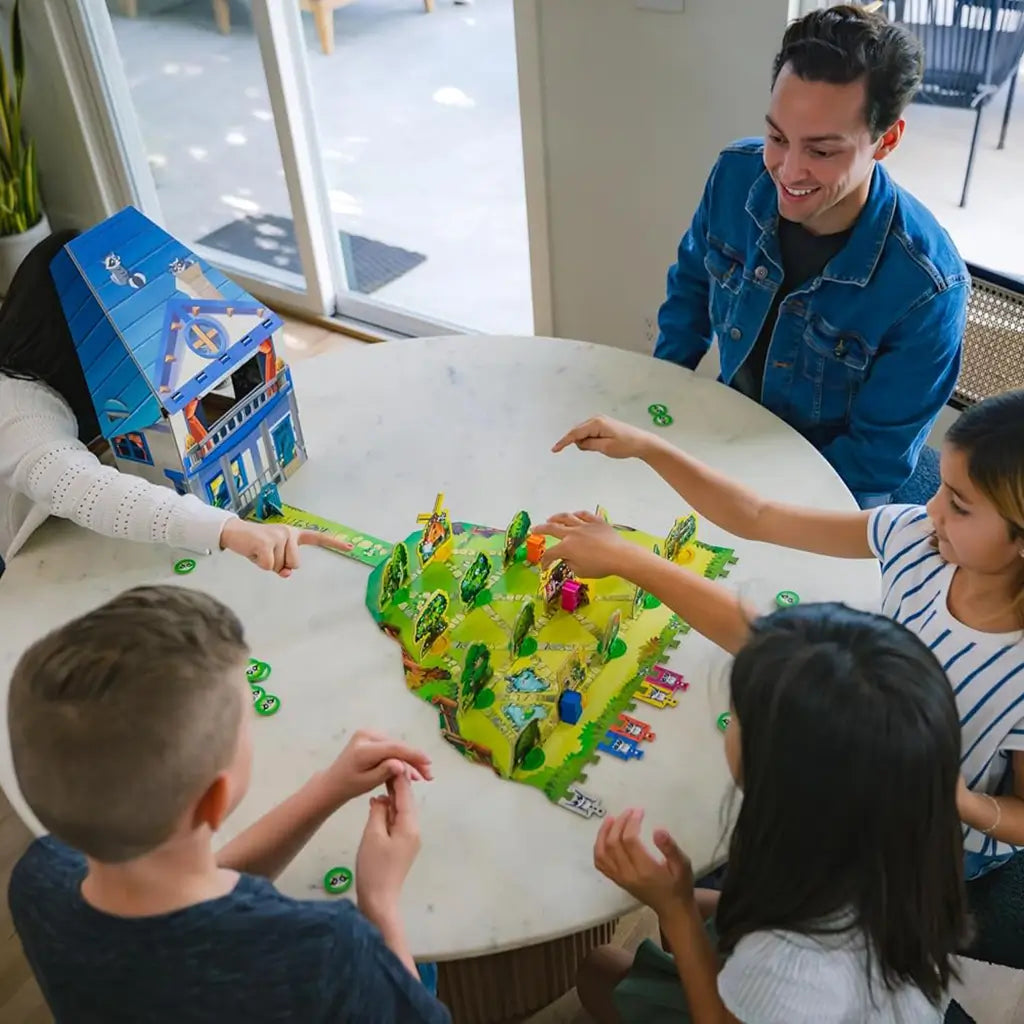 Family of four children and one adult play Ravensburger Garden Heist around a white round table, with colorful 3D garden pieces, raccoon tokens, and a tall blue cardboard house centerpiece.