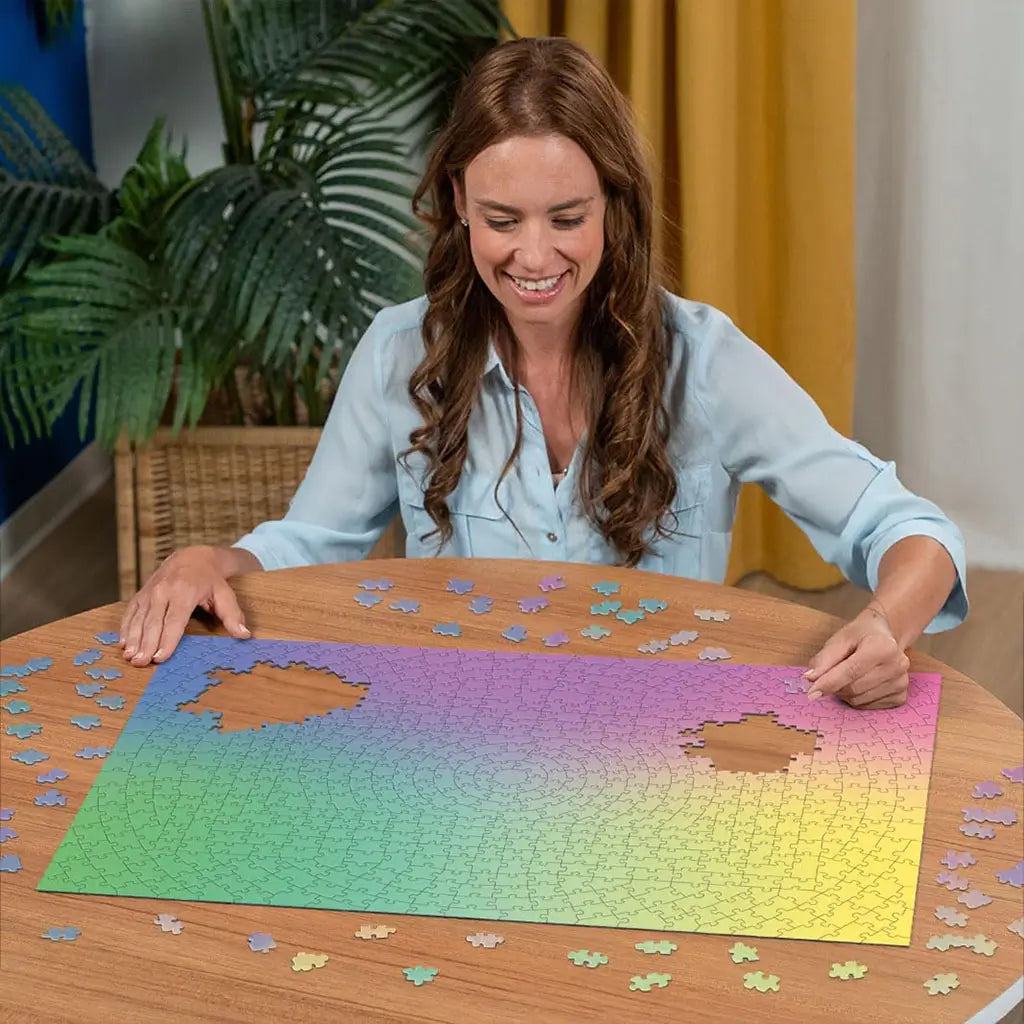 A smiling woman assembles the Ravensburger Krypt gradient puzzle at a round wooden table, surrounded by scattered pastel pieces in a cozy indoor setting.