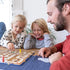 Two young children and an adult happily playing Ravensburger Labyrinth Junior at a wooden table, smiling as they move colorful maze pieces in a family setting.